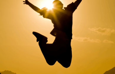 Silhouette of a person jumping against a vibrant sunset. The sun is positioned behind the person, creating a halo effect. Mountains and clouds are visible in the distance. The image conveys a sense of freedom and joy.