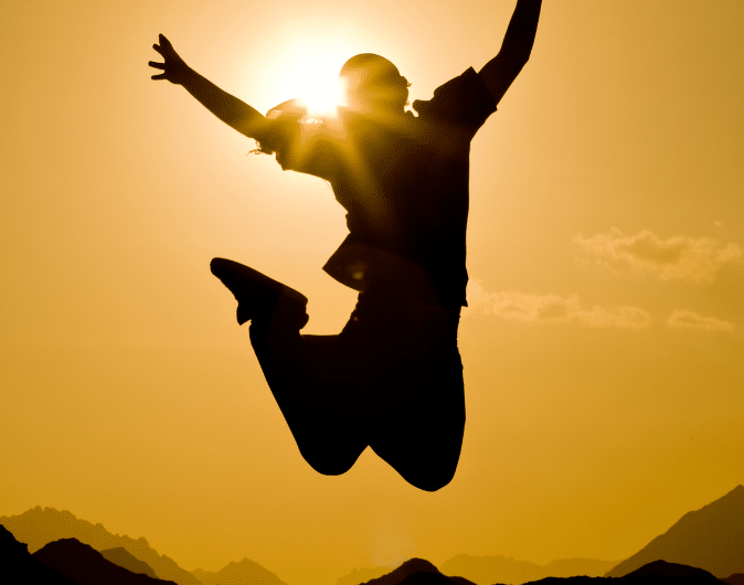 Silhouette of a person jumping against a vibrant sunset. The sun is positioned behind the person, creating a halo effect. Mountains and clouds are visible in the distance. The image conveys a sense of freedom and joy.