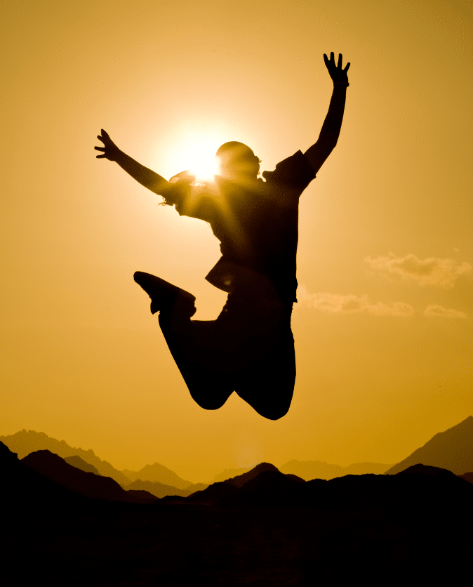 Silhouette of a person jumping against a vibrant sunset. The sun is positioned behind the person, creating a halo effect. Mountains and clouds are visible in the distance. The image conveys a sense of freedom and joy.