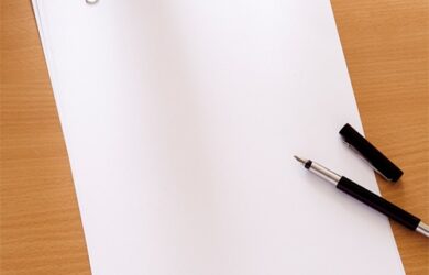 A stack of blank white paper held together with a black binder clip sits on a wooden desk. A black fountain pen with its cap off is placed beside the paper.