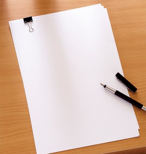 A stack of blank white paper held together with a black binder clip sits on a wooden desk. A black fountain pen with its cap off is placed beside the paper.