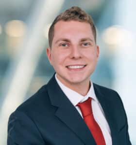 A person in a navy suit and red tie smiles at the camera. The background is softly blurred with a mix of lights and shadows, suggesting an indoor setting.