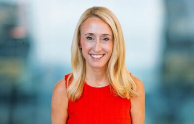 A woman with long blonde hair smiles at the camera. She is wearing a sleeveless red top. The background is softly blurred, consisting of cool blue and gray tones.