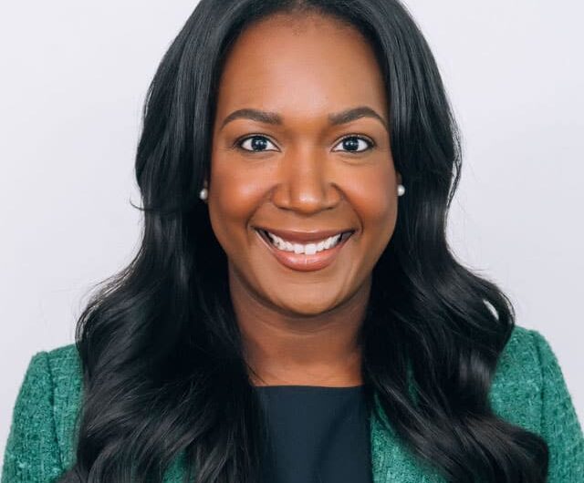 Smiling person with long dark hair wearing a green blazer and black top against a light background.