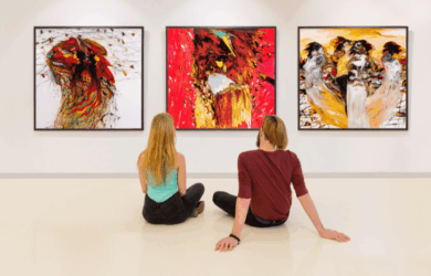 Two people sit on the floor of an art gallery, facing three large, colorful abstract paintings on the wall. The paintings feature vibrant reds, yellows, and contrasting elements. The gallery has a clean, modern aesthetic.