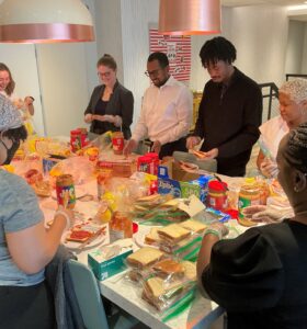 A group of people stands around a table making sandwiches. The table is filled with bread, deli meats, cheese, and sandwich bags. They seem engaged in an organized activity, possibly a community service or group event.