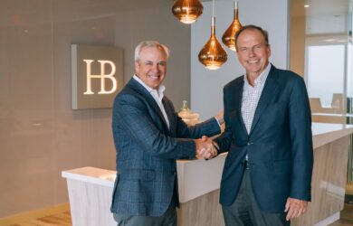 Two men in business attire shake hands in a modern office setting. They stand in front of a reception desk with the initials HB on the wall. Copper-colored pendant lights hang from the ceiling, adding a warm touch to the space.