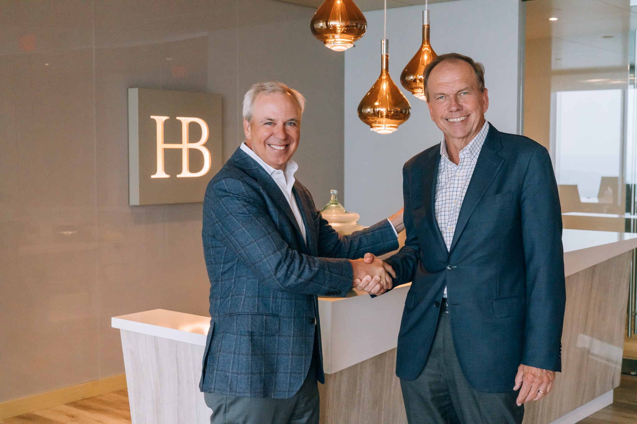 Two men in business attire shake hands in a modern office setting. They stand in front of a reception desk with the initials HB on the wall. Copper-colored pendant lights hang from the ceiling, adding a warm touch to the space.