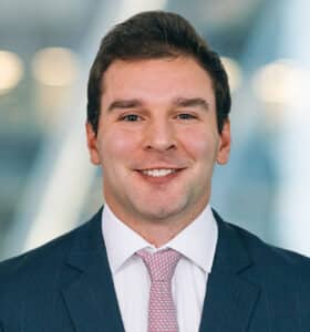 A smiling man in a business suit and pink tie stands against a blurred background with warm lights.