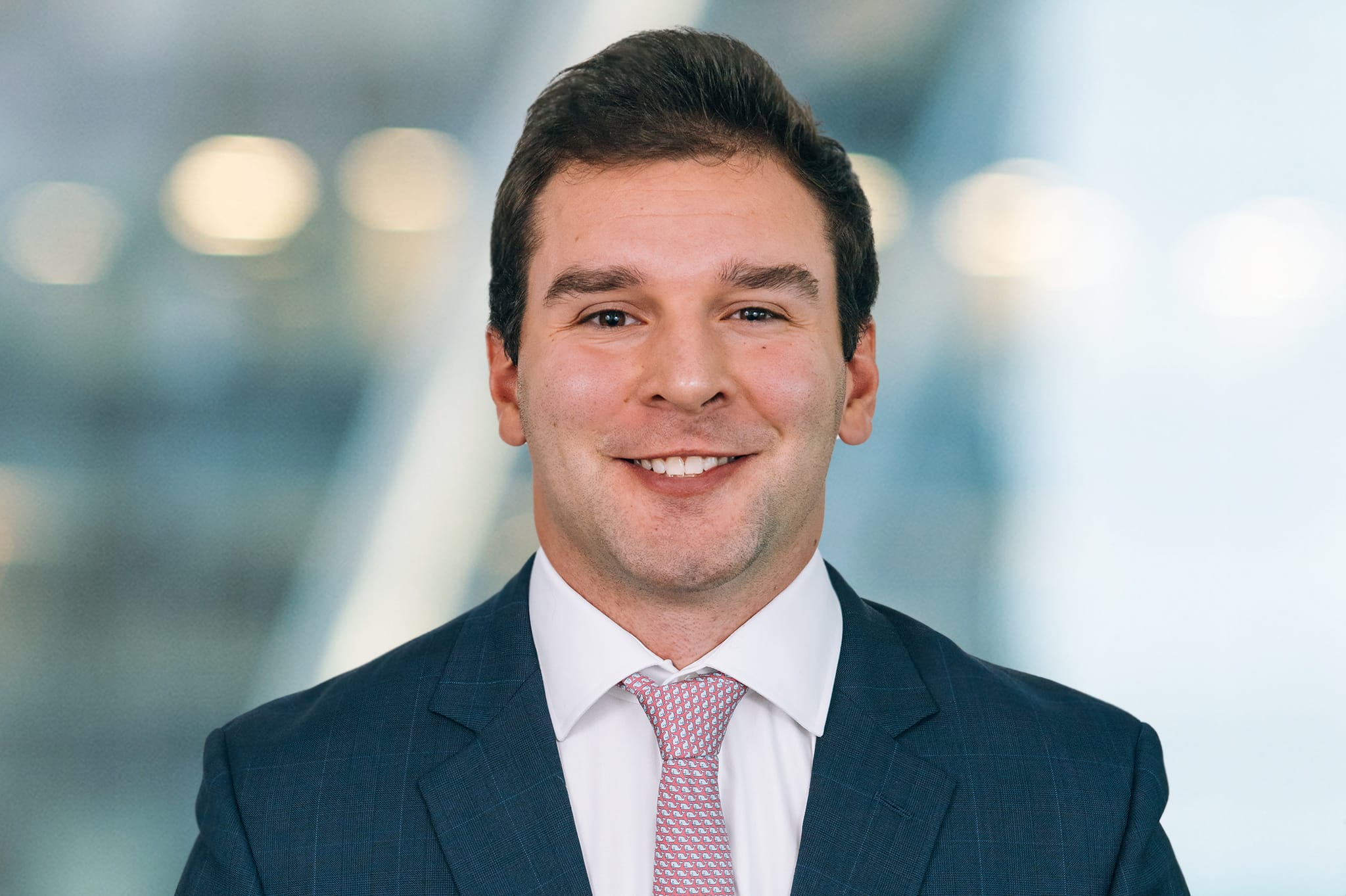 A smiling man in a business suit and pink tie stands against a blurred background with warm lights.