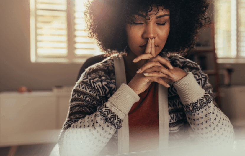 A woman with curly hair sits indoors, wearing a patterned sweater. She rests her chin on her clasped hands, eyes closed, appearing thoughtful or meditative. Soft sunlight filters through window blinds in the background.