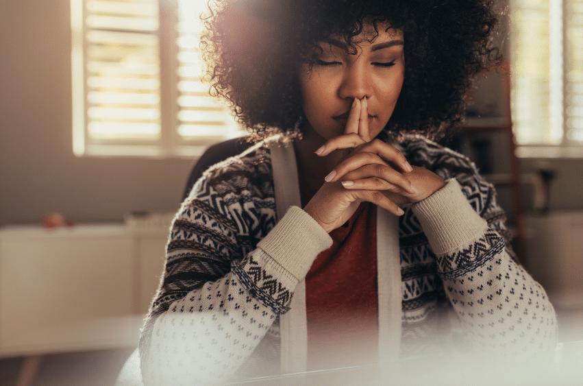 A woman with curly hair sits indoors, wearing a patterned sweater. She rests her chin on her clasped hands, eyes closed, appearing thoughtful or meditative. Soft sunlight filters through window blinds in the background.