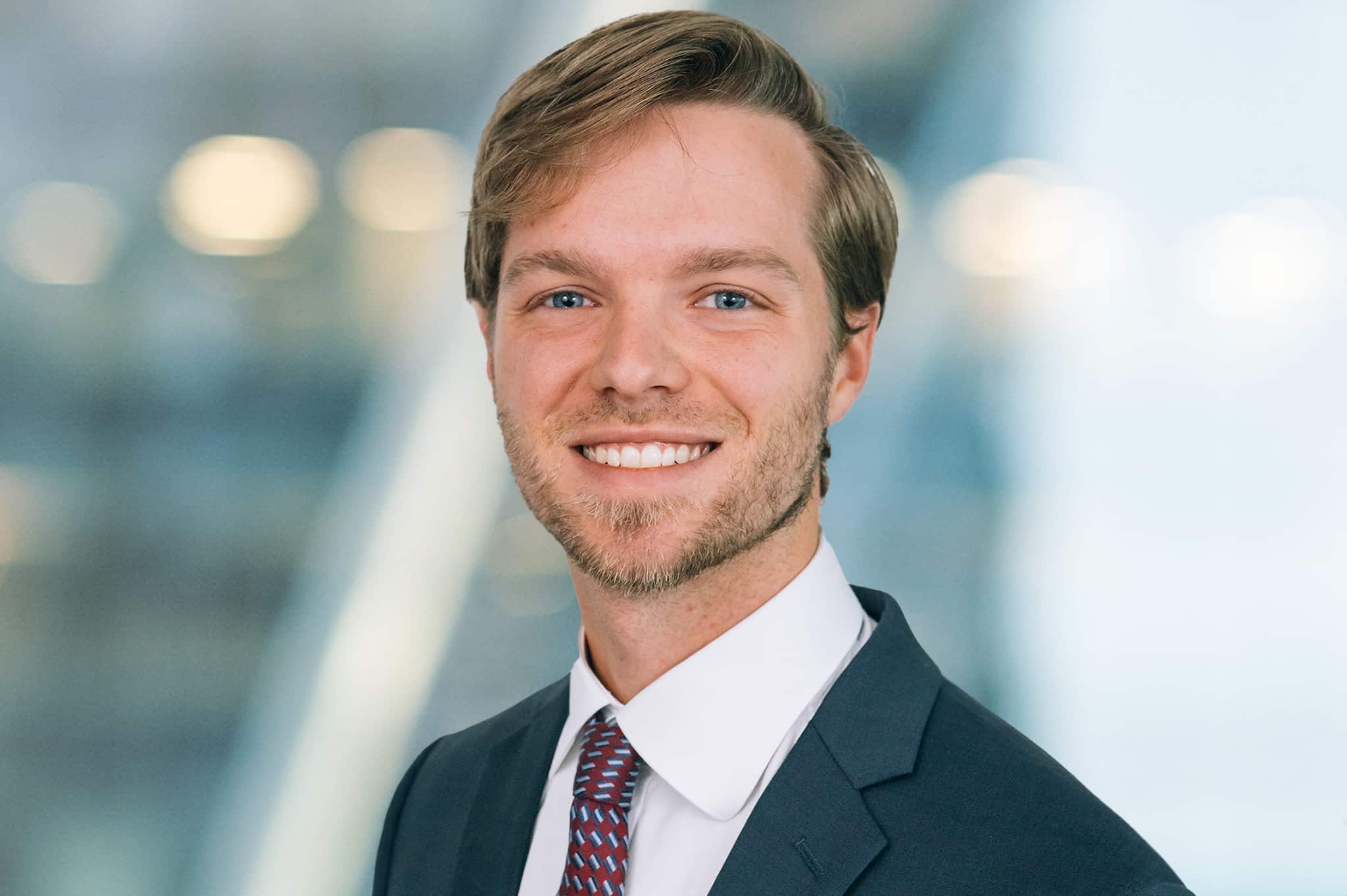 A man in a suit and tie is smiling against a blurred background. He has short, light brown hair and a beard. The setting appears to be a professional or office environment.