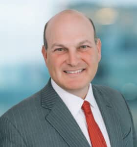 A person in a gray pinstripe suit and red tie is smiling at the camera. The background is blurred, showing an office environment.