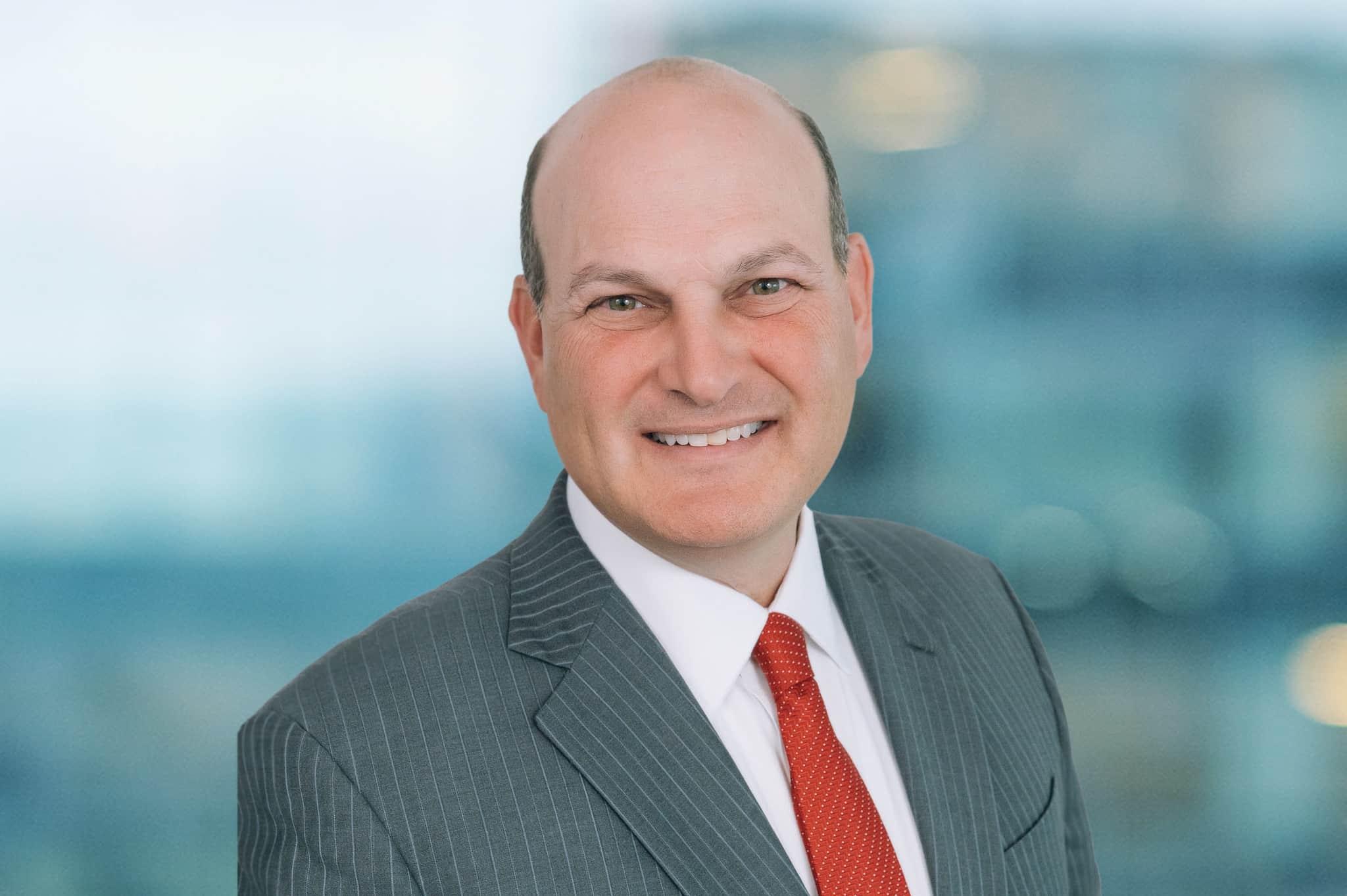 A person in a gray pinstripe suit and red tie is smiling at the camera. The background is blurred, showing an office environment.