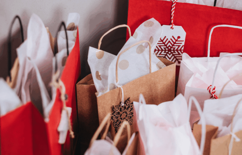A collection of festive gift bags in red and brown, some with tissue paper and decorative tags. The bags are arranged closely together, creating a holiday or celebratory atmosphere.