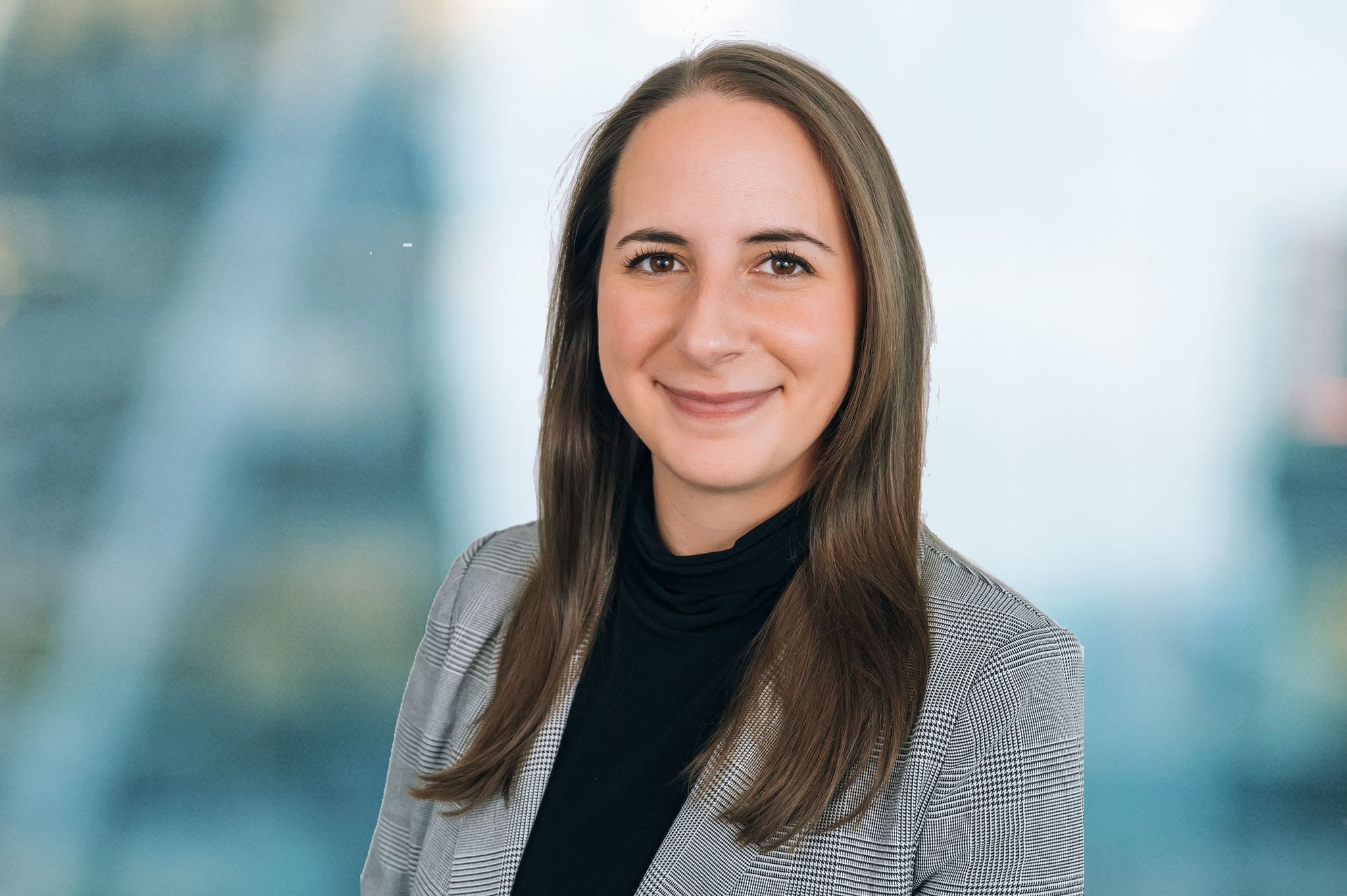 A person with long brown hair is smiling and wearing a gray checkered blazer over a black top. The background is a blurred office setting with large windows.