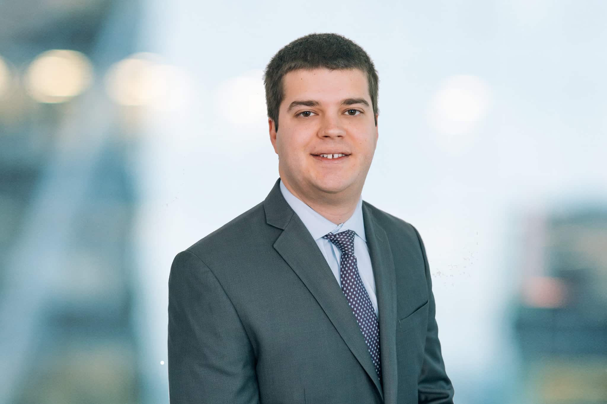A man in a suit and tie stands smiling in front of a blurred background. He has short dark hair and is wearing a gray suit with a light blue shirt and a patterned tie.