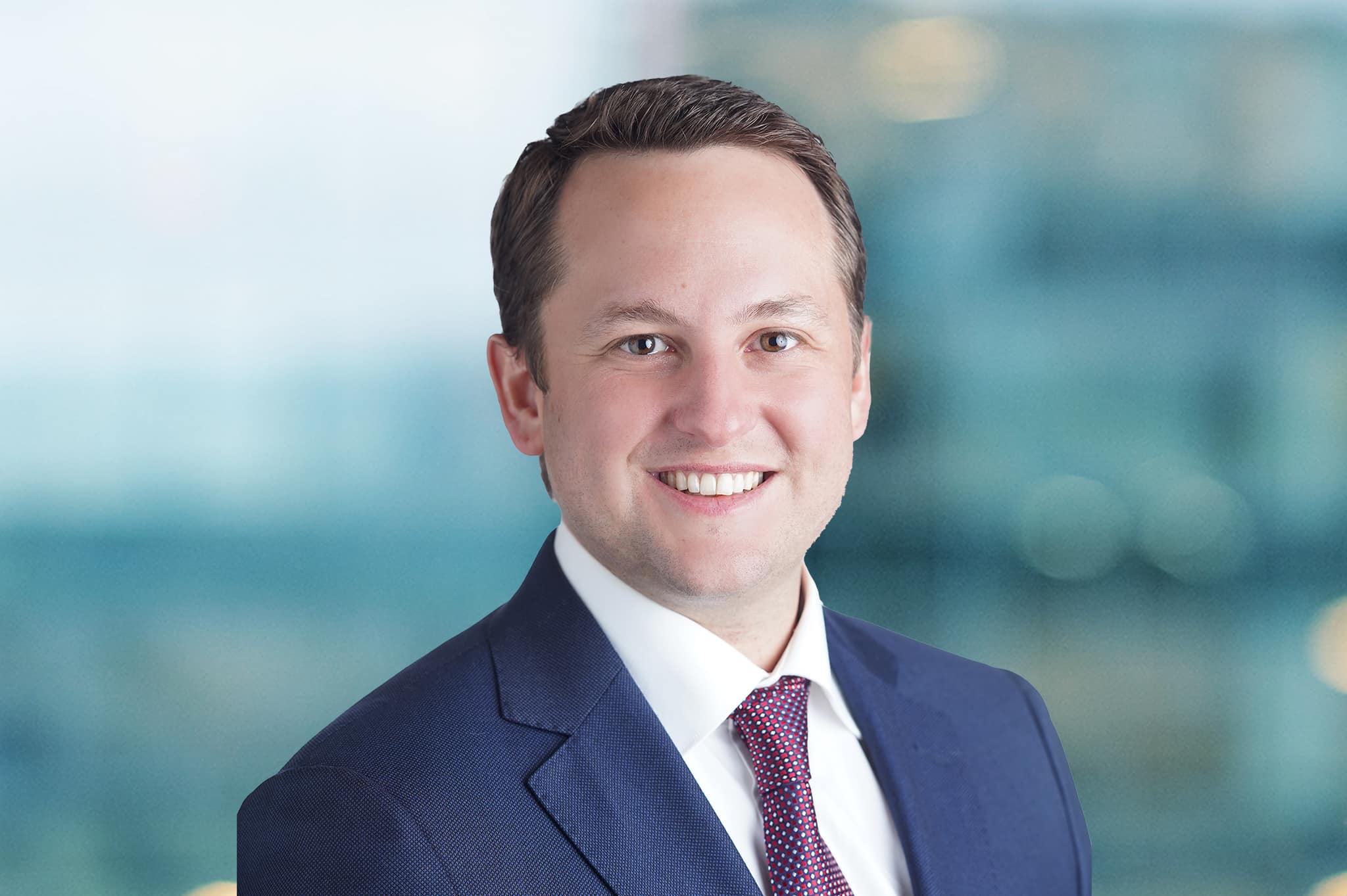 A man in a blue suit and red tie smiles at the camera. The background is a blurred office setting with blue and green tones.