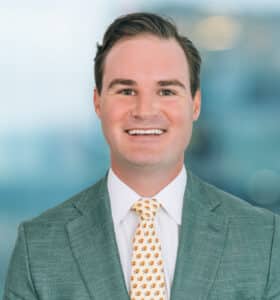 A smiling man with short dark hair is wearing a light green suit jacket, a white dress shirt, and a patterned tie with small orange designs. The background is a blurred cityscape.