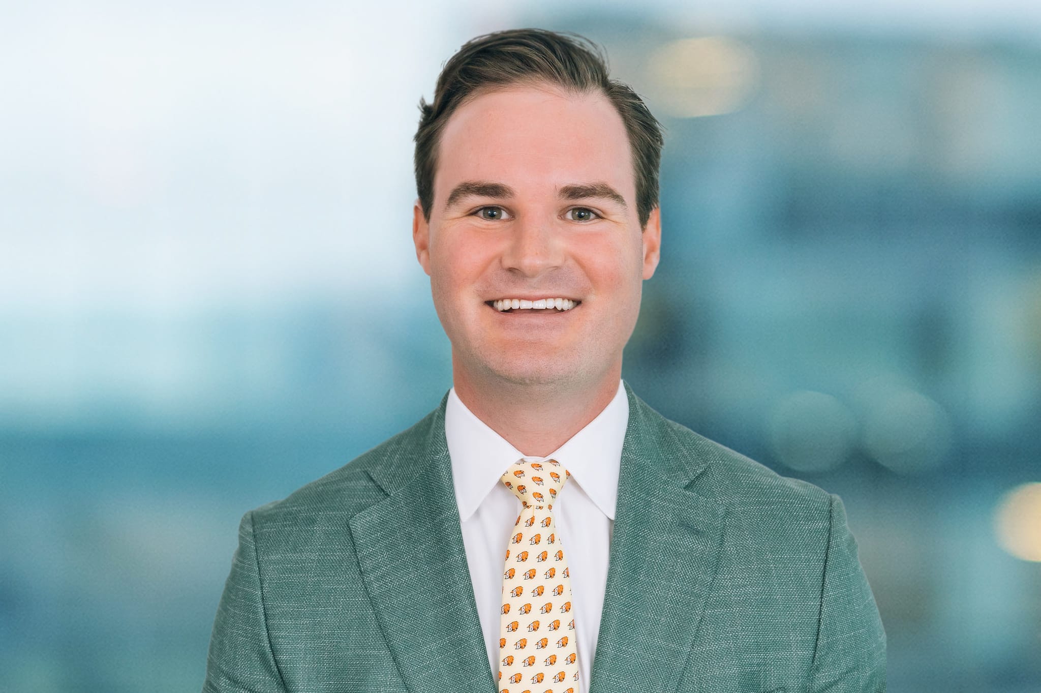 A smiling man with short dark hair is wearing a light green suit jacket, a white dress shirt, and a patterned tie with small orange designs. The background is a blurred cityscape.