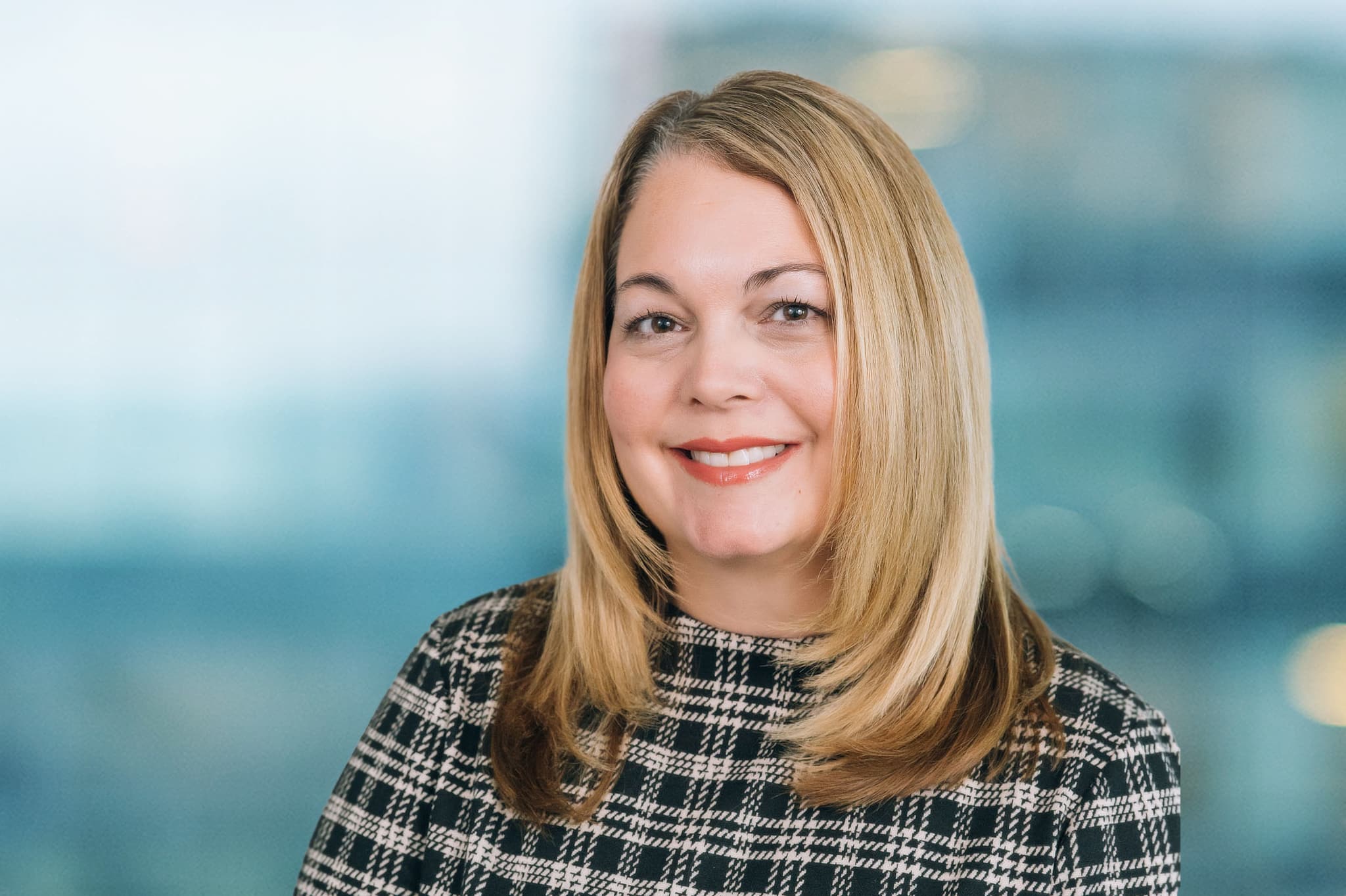 A woman with long blonde hair smiles while wearing a black and white patterned top. She stands in front of a blurred background that suggests an office environment.