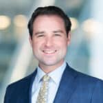 A man in a blue suit and patterned tie smiles at the camera. The blurred background shows circular lights and a softly colored wall, suggesting a professional setting.