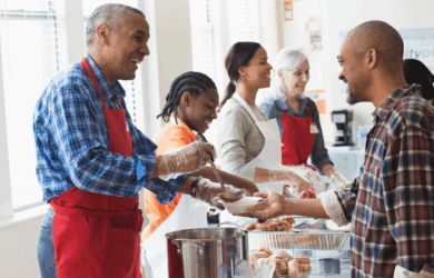 A diverse group of people at a community kitchen serve meals to individuals in line. Smiles and friendly interactions are visible as volunteers wearing red aprons distribute food. Natural light fills the room through nearby windows.