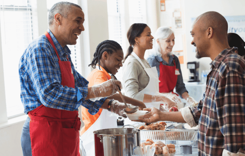 A diverse group of people at a community kitchen serve meals to individuals in line. Smiles and friendly interactions are visible as volunteers wearing red aprons distribute food. Natural light fills the room through nearby windows.