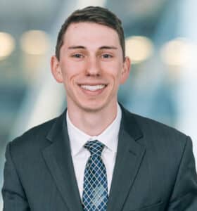 A smiling person with short hair is wearing a dark suit, white shirt, and patterned tie. The background is blurred, featuring soft lights.
