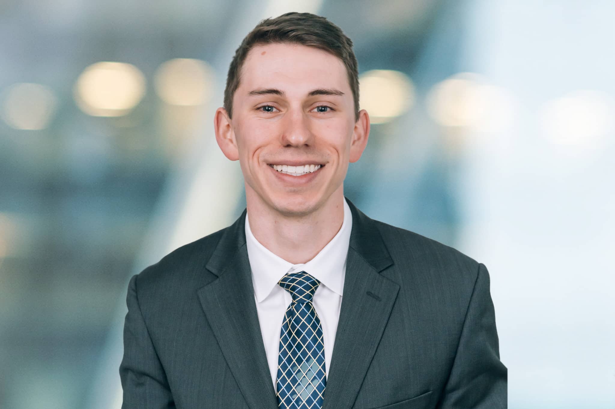 A smiling person with short hair is wearing a dark suit, white shirt, and patterned tie. The background is blurred, featuring soft lights.