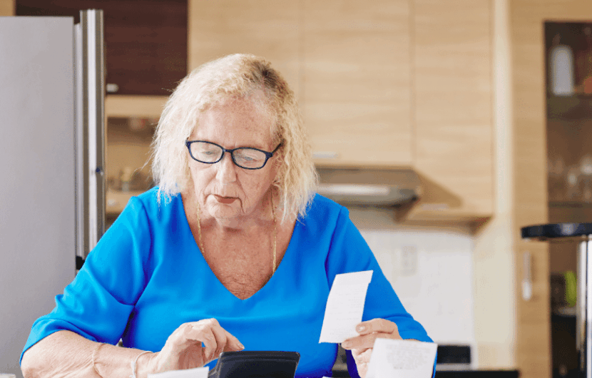 An elderly woman with glasses, wearing a blue shirt, sits at a table in a kitchen. She is looking at papers and a receipt in her hands, appearing focused.