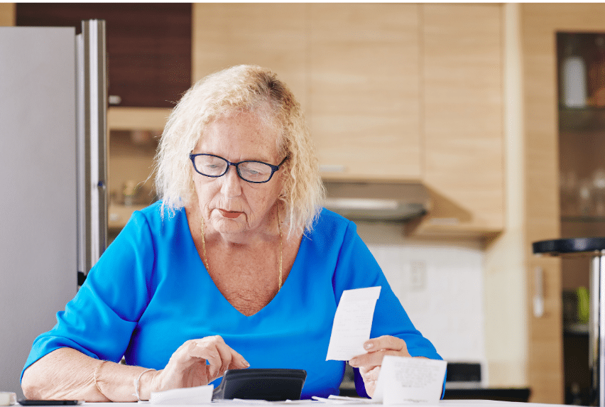 An elderly woman with glasses, wearing a blue shirt, sits at a table in a kitchen. She is looking at papers and a receipt in her hands, appearing focused.