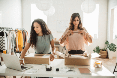Two women work at a table in a clothing store, packing boxes for shipping. One uses a laptop, while the other seals a box. Clothing racks are in the background, and the room is well-lit with paper lanterns hanging from the ceiling.