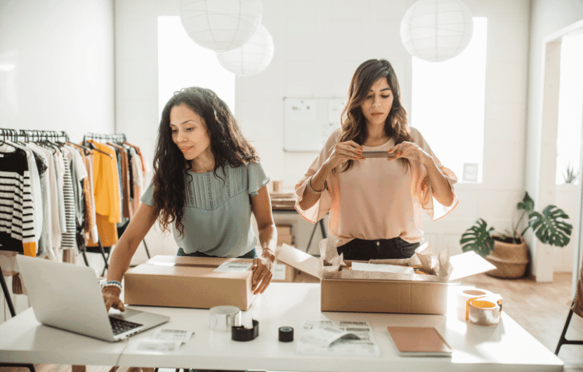 Two women work at a table in a clothing store, packing boxes for shipping. One uses a laptop, while the other seals a box. Clothing racks are in the background, and the room is well-lit with paper lanterns hanging from the ceiling.