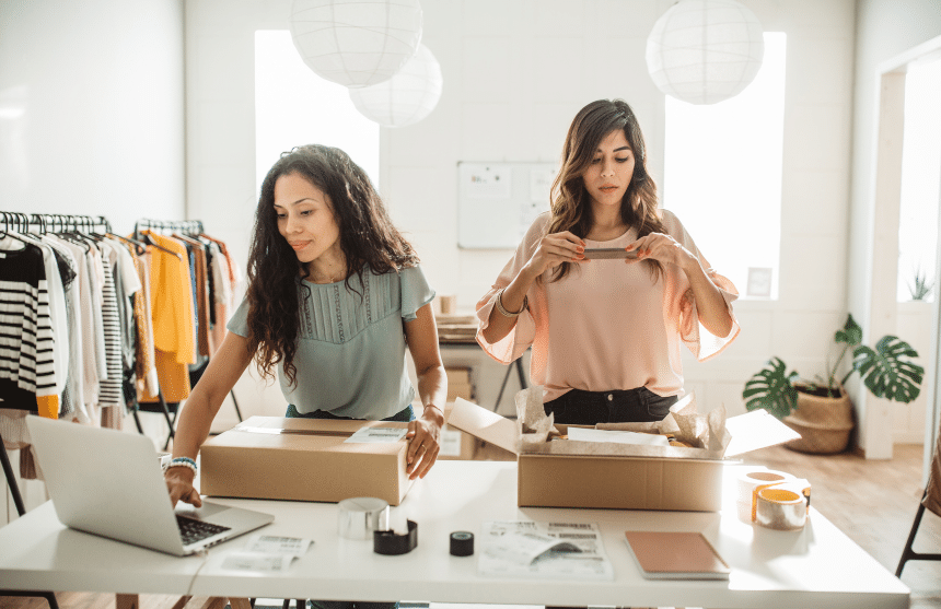 Two women work at a table in a clothing store, packing boxes for shipping. One uses a laptop, while the other seals a box. Clothing racks are in the background, and the room is well-lit with paper lanterns hanging from the ceiling.
