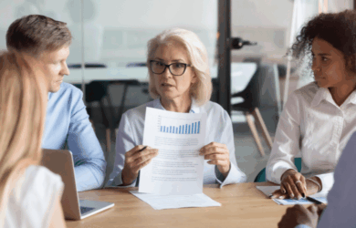 A group of four diverse professionals in a meeting room, focused on a senior woman holding documents with charts. She appears to be explaining data to the others who are attentively listening. A laptop is visible on the table.