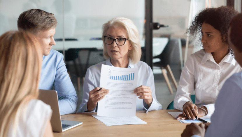 A group of four diverse professionals in a meeting room, focused on a senior woman holding documents with charts. She appears to be explaining data to the others who are attentively listening. A laptop is visible on the table.