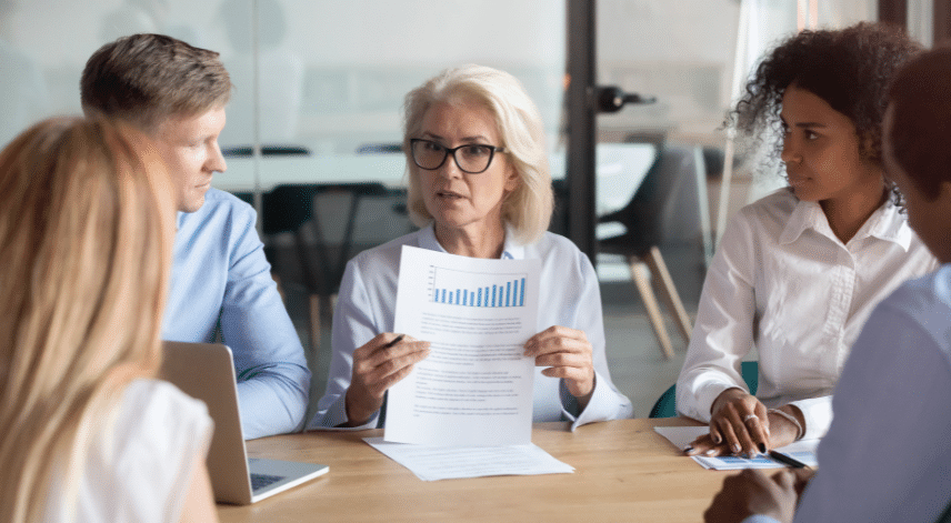 A group of four diverse professionals in a meeting room, focused on a senior woman holding documents with charts. She appears to be explaining data to the others who are attentively listening. A laptop is visible on the table.