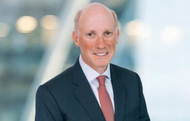 A man with a bald head, wearing a dark suit, white shirt, and red tie, smiles at the camera. The background is blurred with soft lighting.