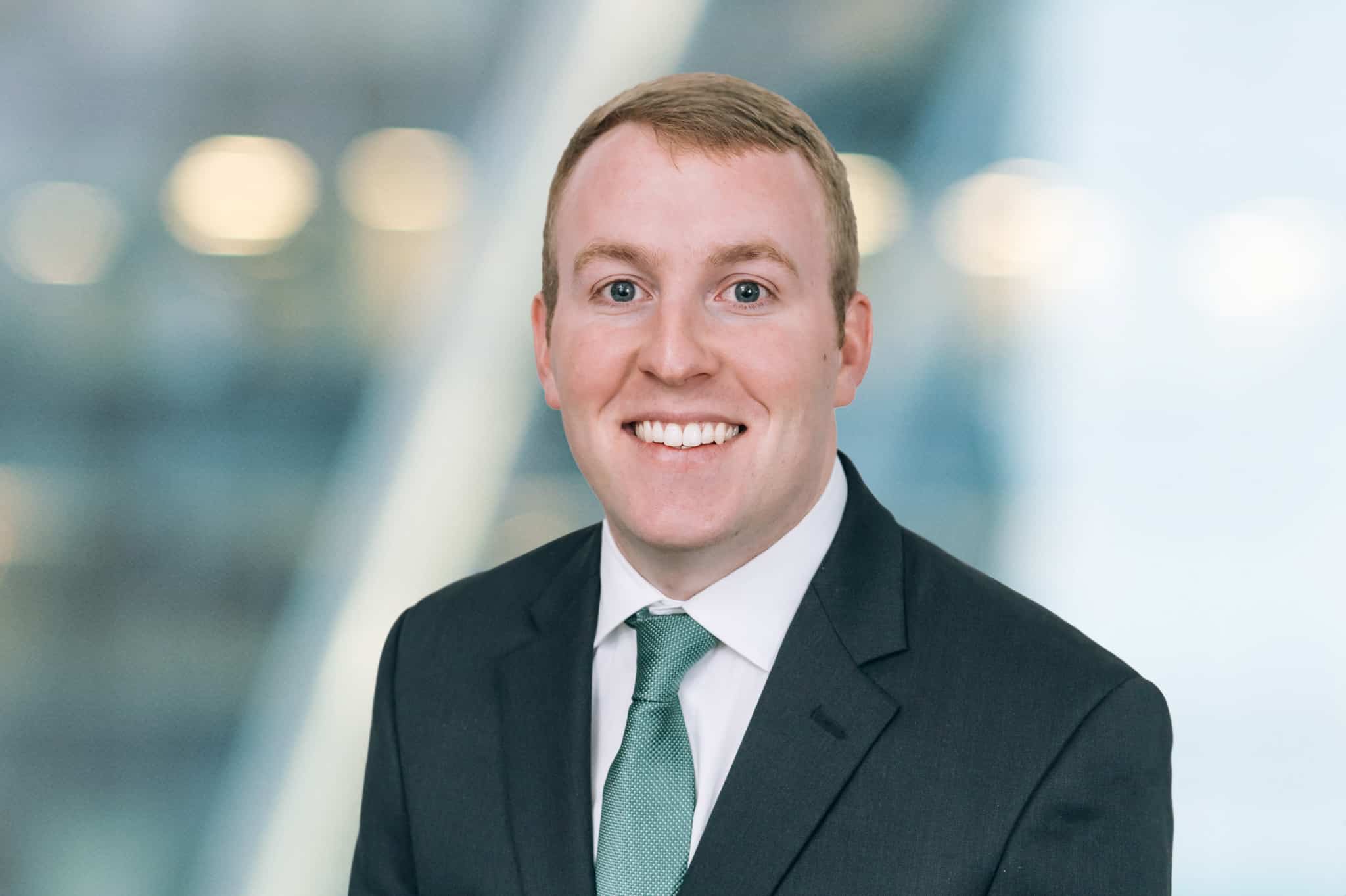 A smiling man in a suit with a green tie poses against a blurred background.