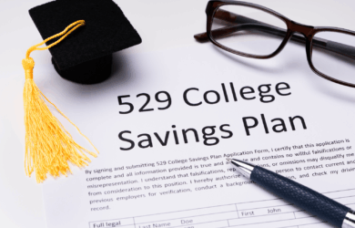 A 529 College Savings Plan application form is shown. A small graduation cap is placed on the top left, with a pair of glasses and a pen nearby on a white surface.