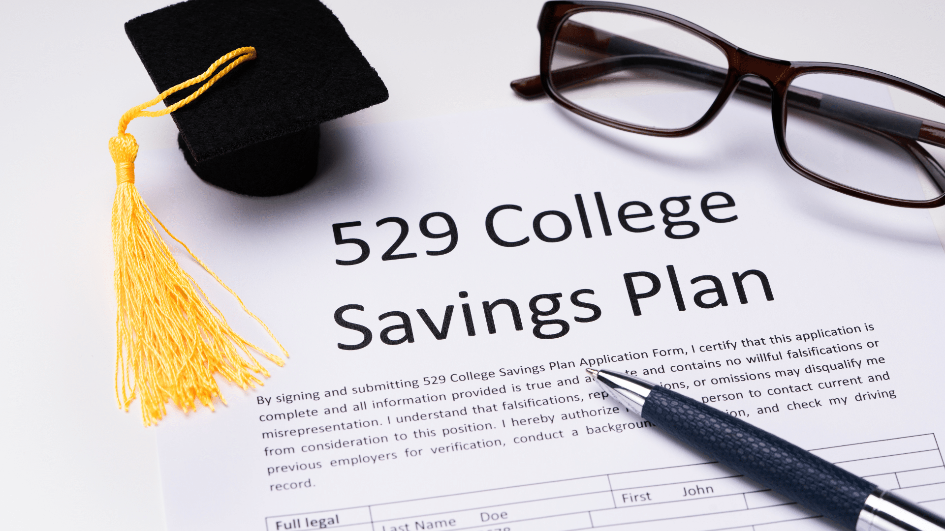 A 529 College Savings Plan application form is shown. A small graduation cap is placed on the top left, with a pair of glasses and a pen nearby on a white surface.