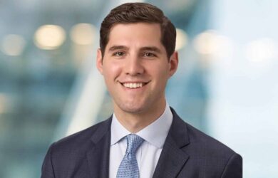 A young man with short dark hair smiles while wearing a dark suit, white shirt, and light blue tie. He stands against a blurred blue and white background, giving a professional and approachable appearance.