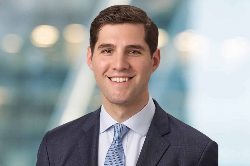 A young man with short dark hair smiles while wearing a dark suit, white shirt, and light blue tie. He stands against a blurred blue and white background, giving a professional and approachable appearance.