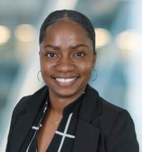 A smiling person with a neat hairstyle wearing a black blazer and a checkered shirt. The background is blurred with soft lighting, creating a professional and approachable appearance.