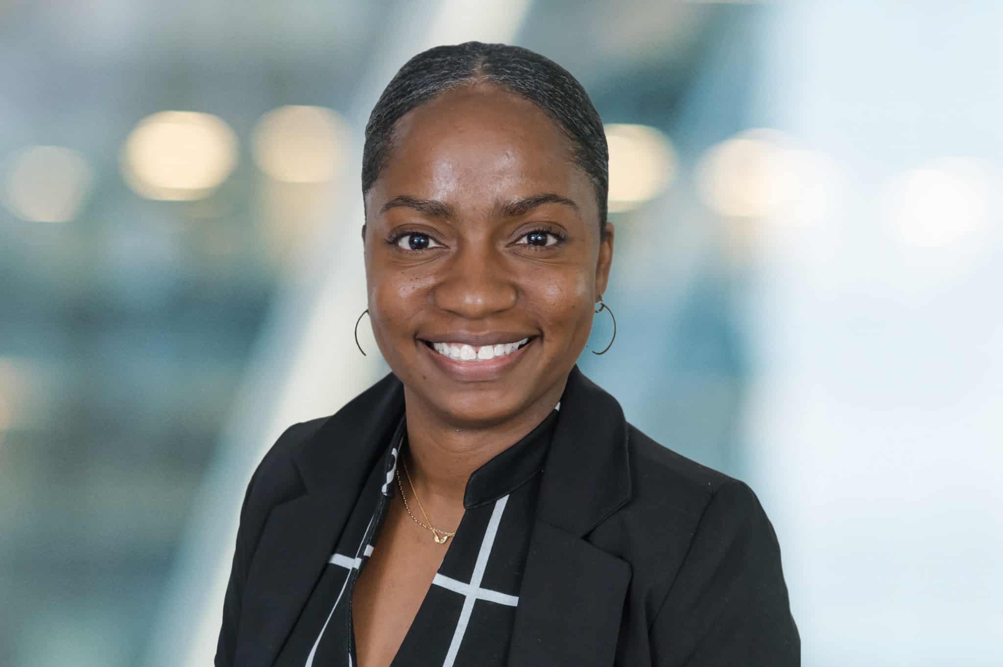 A smiling person with a neat hairstyle wearing a black blazer and a checkered shirt. The background is blurred with soft lighting, creating a professional and approachable appearance.