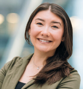 A woman with long brown hair smiles confidently at the camera, wearing a green blazer over a black top. The background is softly blurred with cool tones and round lights.
