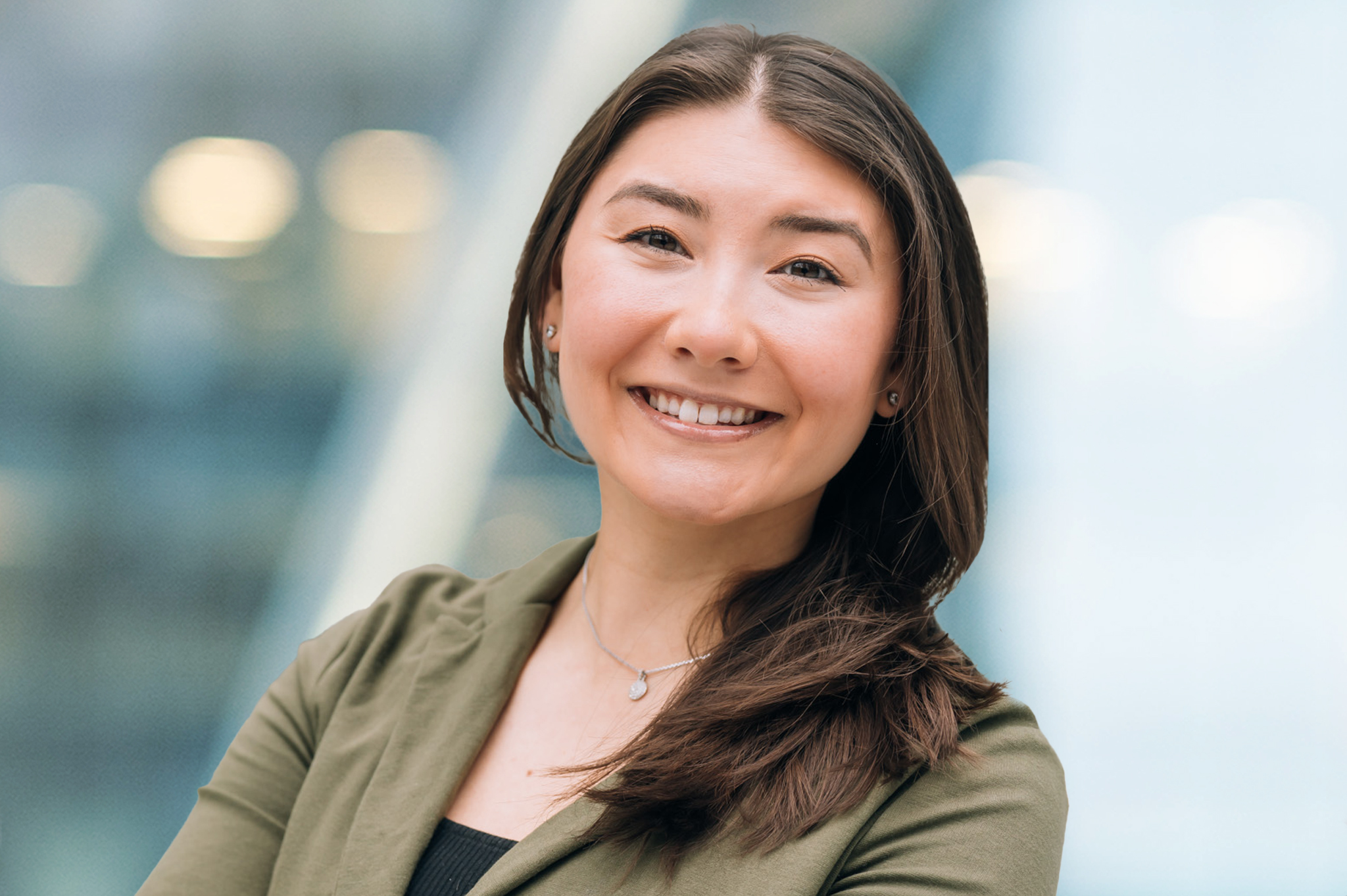 A woman with long brown hair smiles confidently at the camera, wearing a green blazer over a black top. The background is softly blurred with cool tones and round lights.
