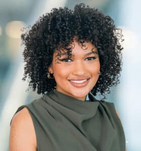 A person with curly hair smiles at the camera, wearing a sleeveless green top. The background is softly blurred with bright lights.